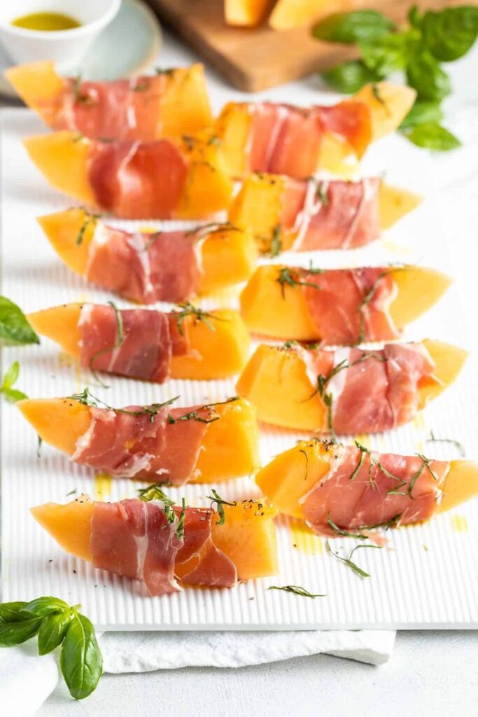 Slices of cantaloupe wrapped in prosciutto garnished with herbs on a white platter for a fresh appetizer.
