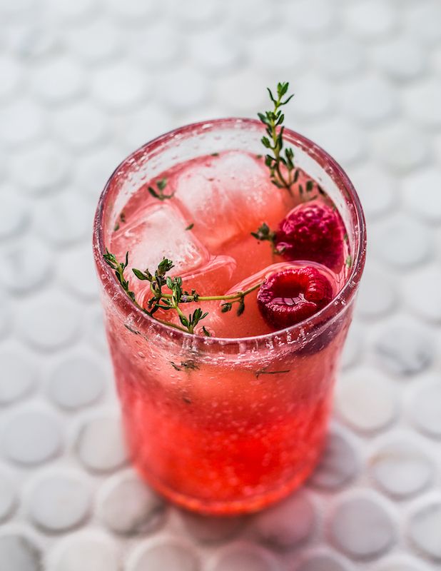 Refreshing raspberry cocktail with ice and thyme sprigs in a tall glass on a light background.