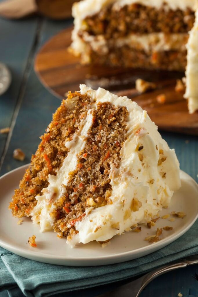 Slice of homemade carrot cake with creamy frosting and walnut topping on a plate.