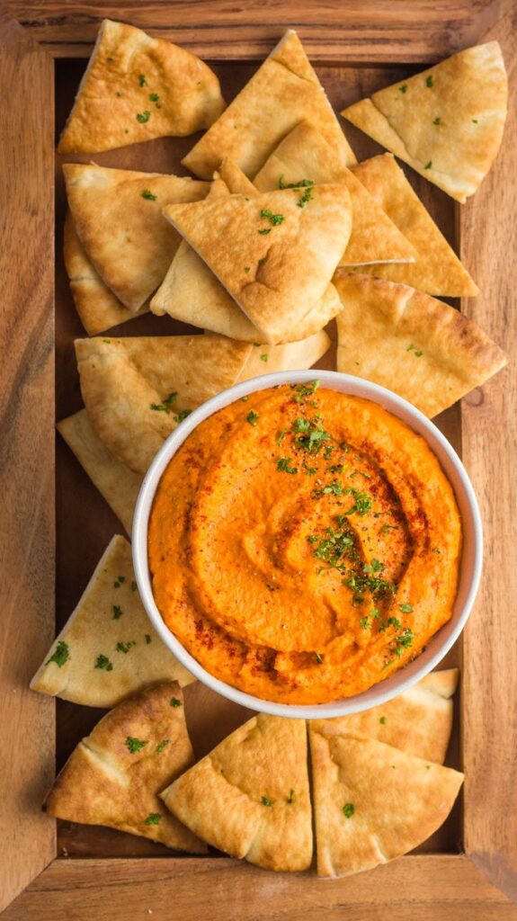 Creamy red pepper hummus with pita chips on a wooden tray, garnished with parsley.