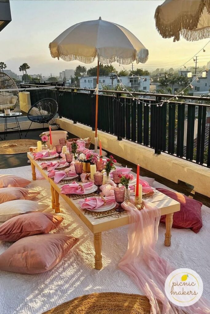 Elegant rooftop picnic setup with pink decor, fringe umbrella, and cozy cushions at sunset.