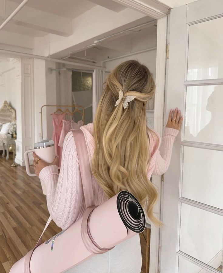 Woman with yoga mat and drink, wearing pastel pink sweater, standing in a bright room with wooden floor.