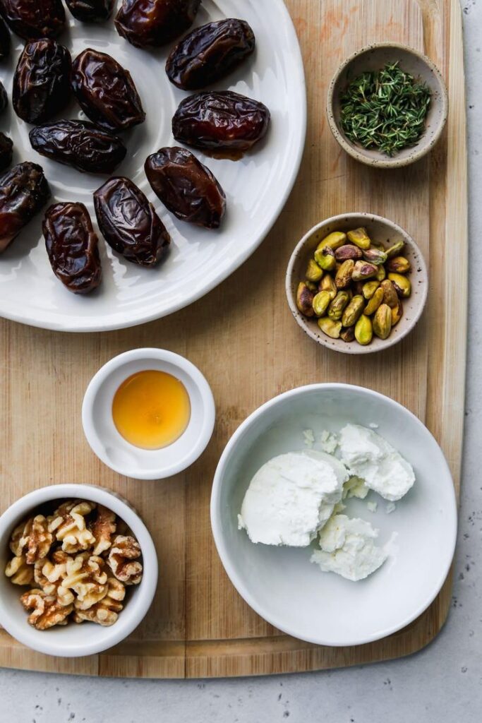 Plate of dates with bowls of goat cheese, walnuts, honey, pistachios, and herbs on wooden board.