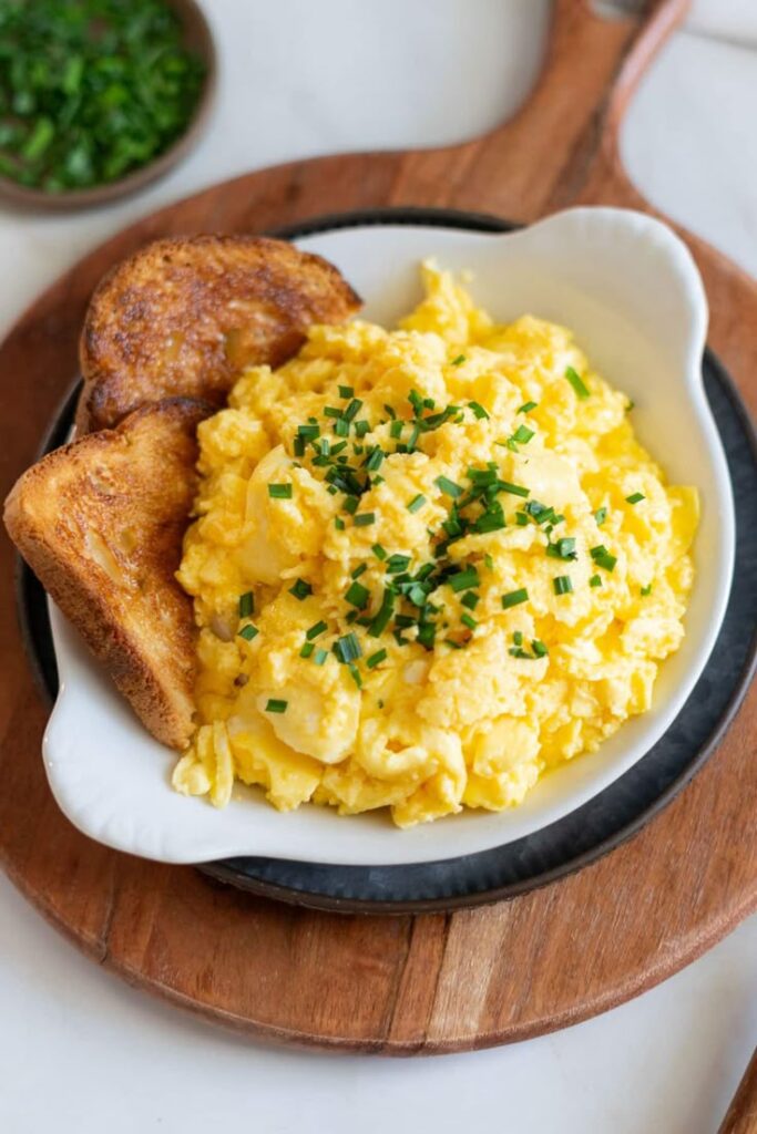 Creamy scrambled eggs with chives and crispy toast on a white plate on a wooden board, perfect breakfast meal.