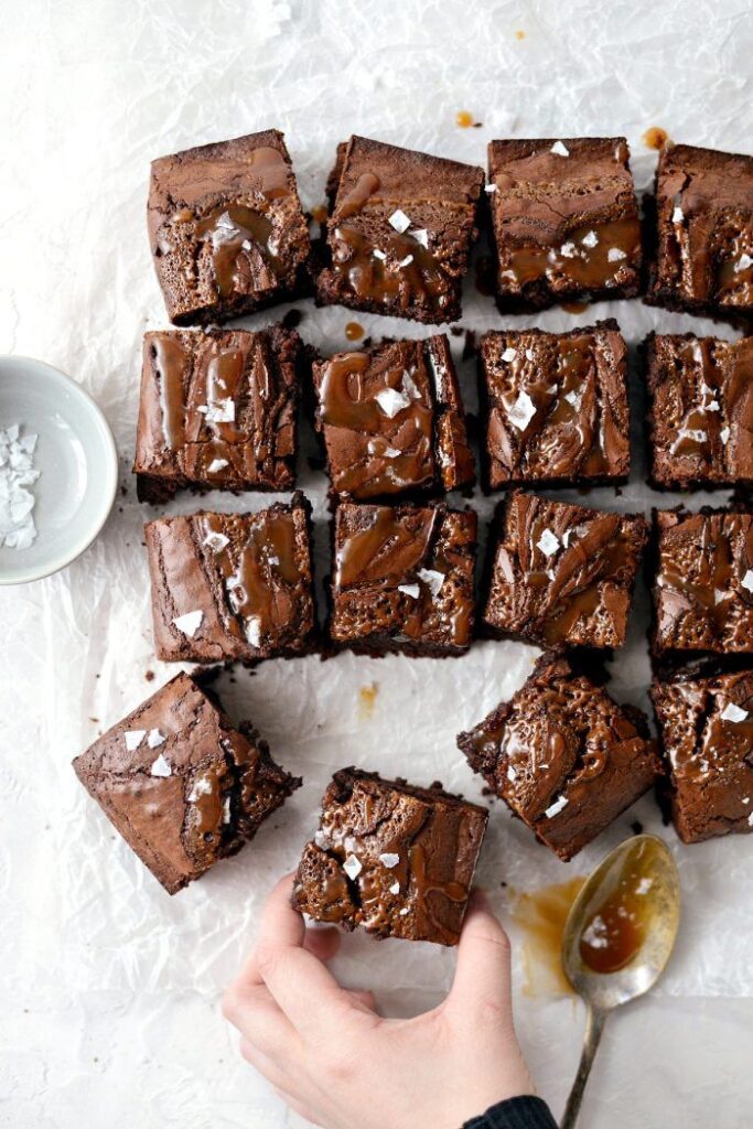 Delicious homemade brownies topped with caramel and sea salt on parchment paper, hand taking a piece.
