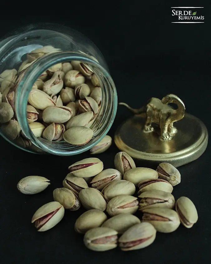 Pistachios spilling from a glass jar with an elephant lid on a dark background.