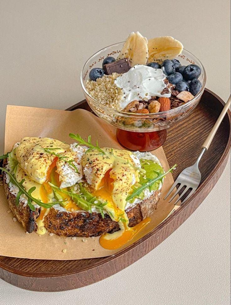 Avocado toast with poached eggs and a yogurt granola bowl with fruits on a wooden tray.