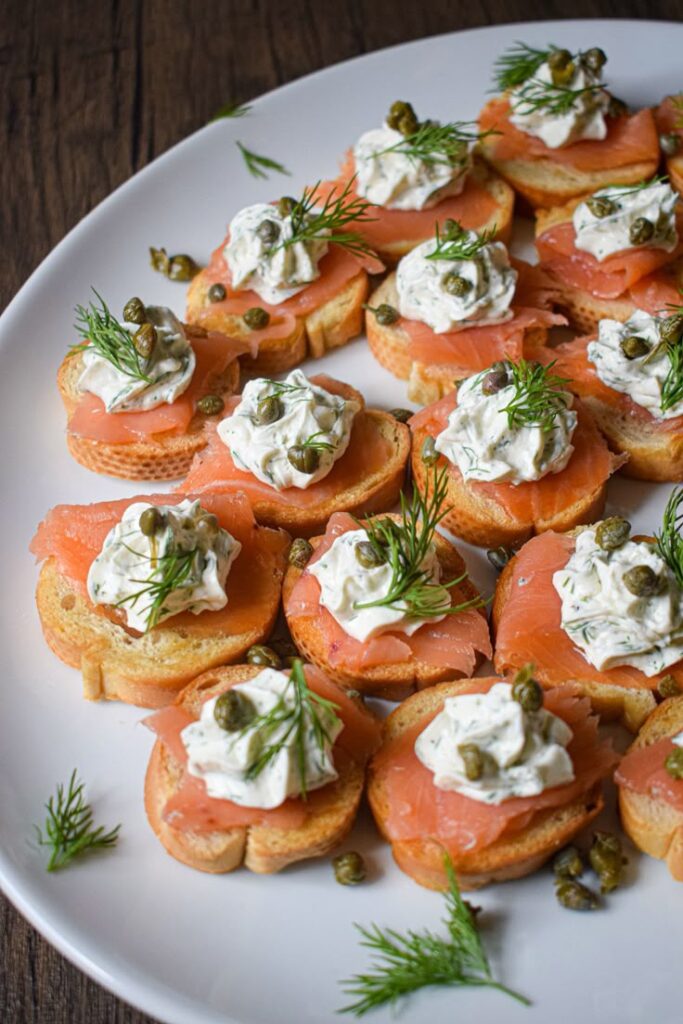 Smoked salmon appetizers with cream cheese, capers, and dill on toasted bread slices on a white platter.