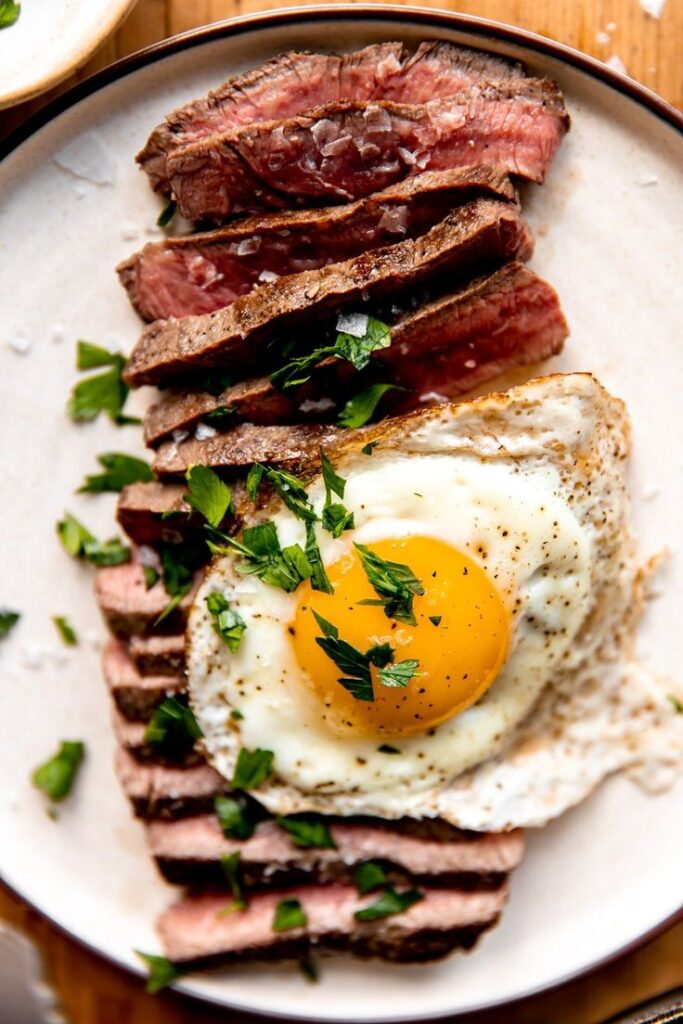 Sliced steak topped with a sunny-side-up egg and fresh herbs on a plate, perfect for a gourmet breakfast.