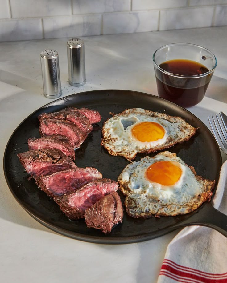 Sizzling steak slices and sunny-side-up eggs on a pan, with a cup of coffee and salt shakers on the side.