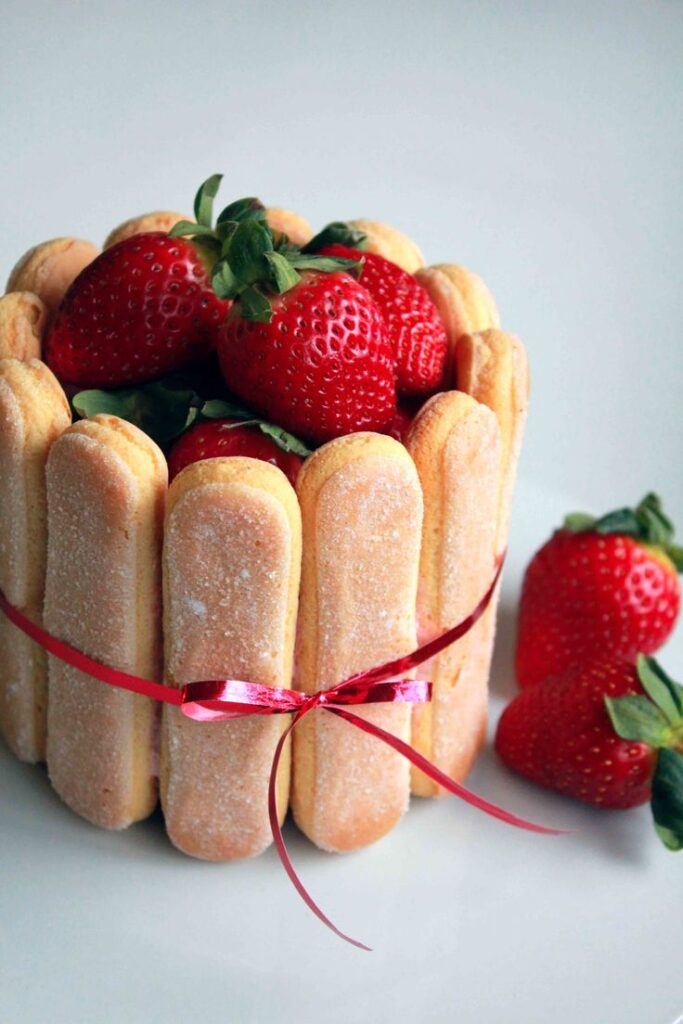 Strawberry charlotte cake with ladyfingers, tied with a red ribbon, garnished with fresh strawberries on top.