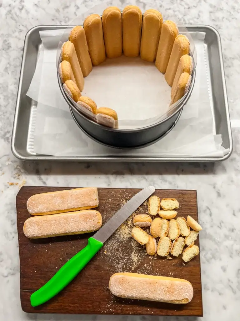 Ladyfingers arranged in a cake pan for charlotte dessert, with a serrated knife and cut pieces on a wooden board.