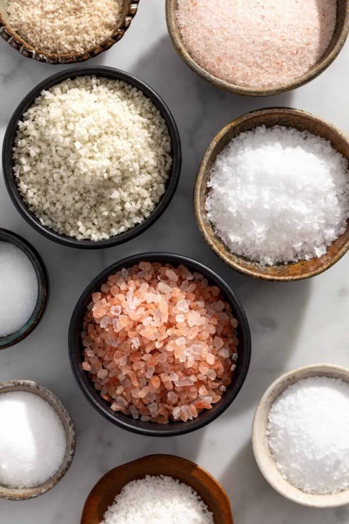 Assorted salts in bowls on marble surface, including Himalayan pink, sea, and kosher varieties.