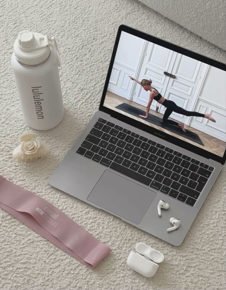 Laptop showing online workout, next to water bottle, resistance band, hair clip, and AirPods on a carpet.
