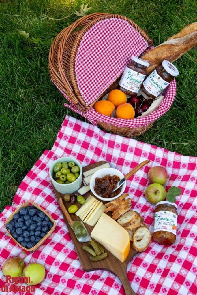 Picnic setup with cheese, olives, bread, jam, fruits, and a wicker basket on a red checkered cloth on grass.