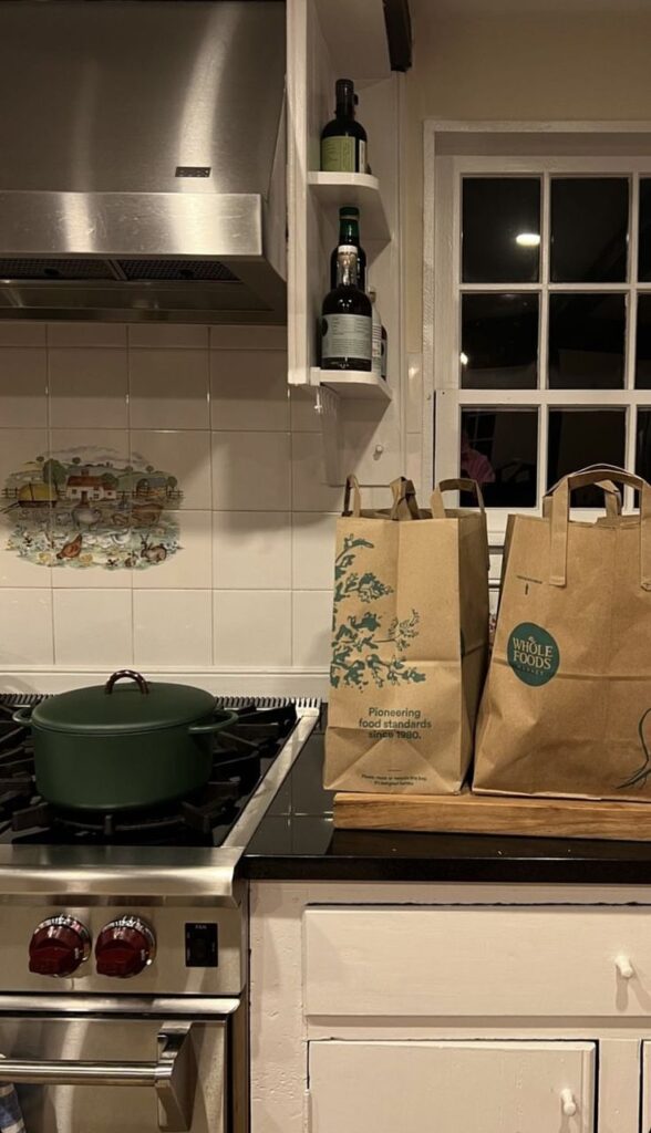 Kitchen with stove, green pot, paper grocery bags, and shelf with olive oil bottles near a window.