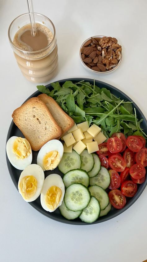 Healthy breakfast plate with boiled eggs, toast, cheese, arugula, cucumbers, cherry tomatoes, nuts, and iced coffee drink.