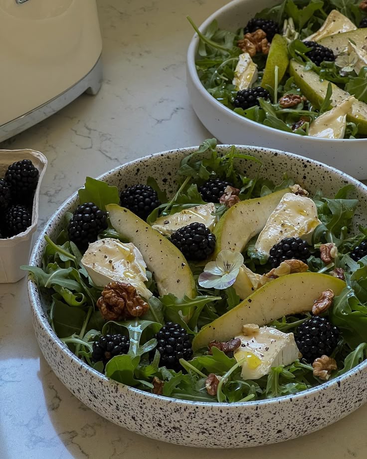 Salad bowl with arugula, pear slices, brie, blackberries, and walnuts on a countertop.