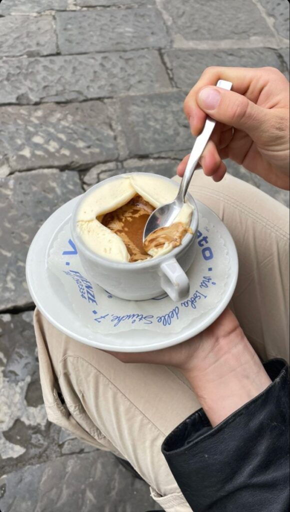 Person enjoying a creamy espresso in a white cup, sitting on cobblestone, holding a spoon.