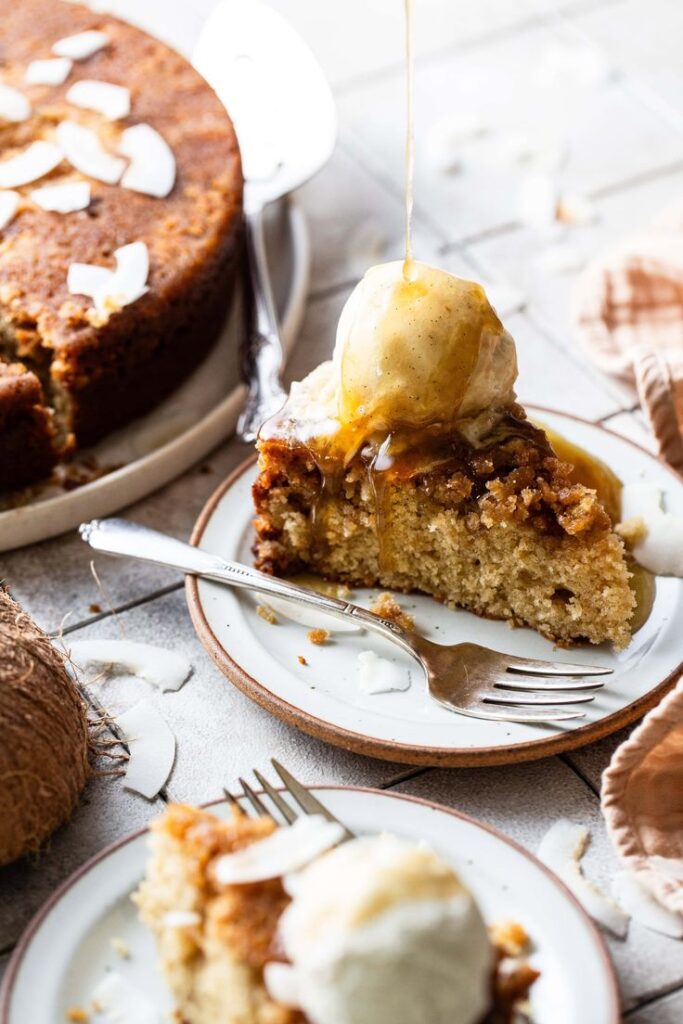 Slice of moist cake topped with ice cream and syrup drizzle on a white plate with coconut flakes.
