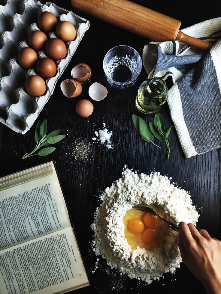 Top-down view of baking ingredients: eggs, flour, oil, and an open cookbook on a dark surface with a rolling pin.
