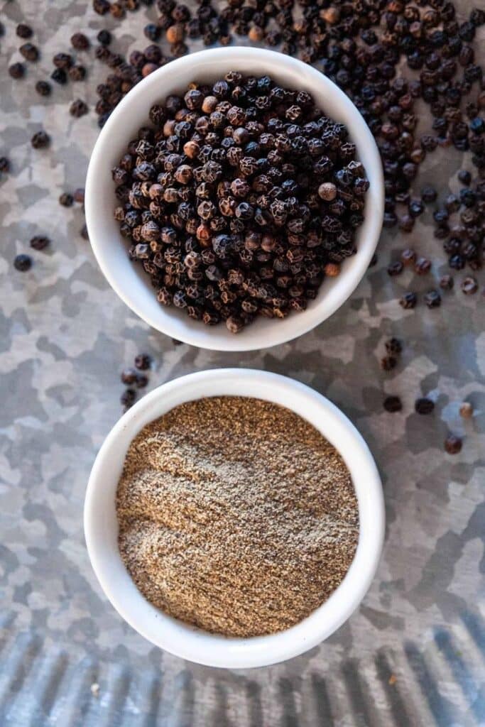 Whole and ground black pepper in white bowls on a metallic surface, highlighting spice variety.