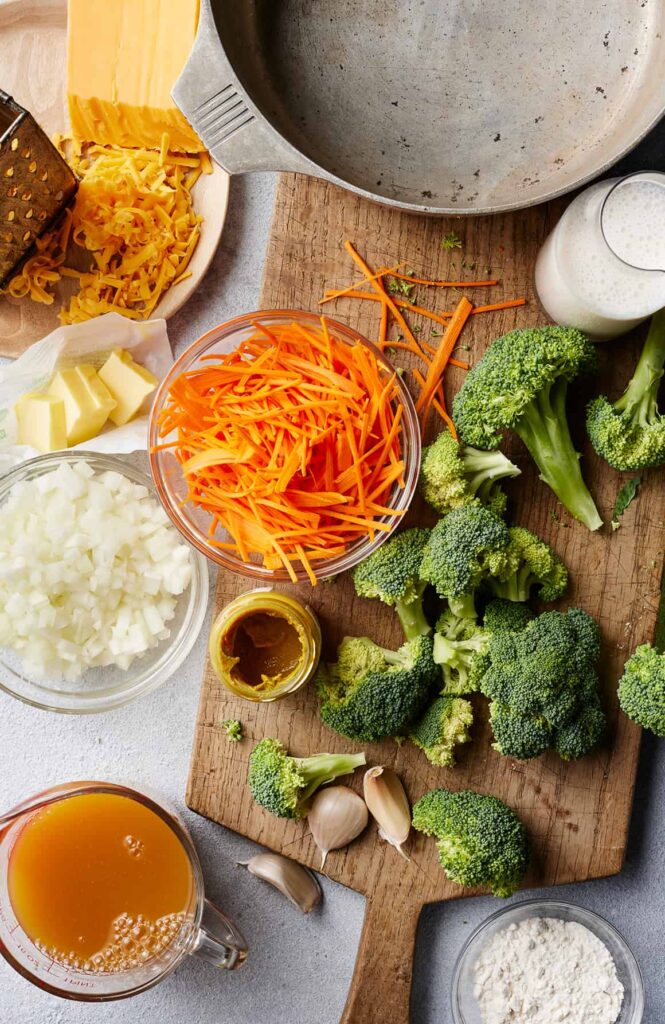 Ingredients for making broccoli cheddar soup: cheese, carrots, broccoli, onions, broth, and milk on a wooden board.