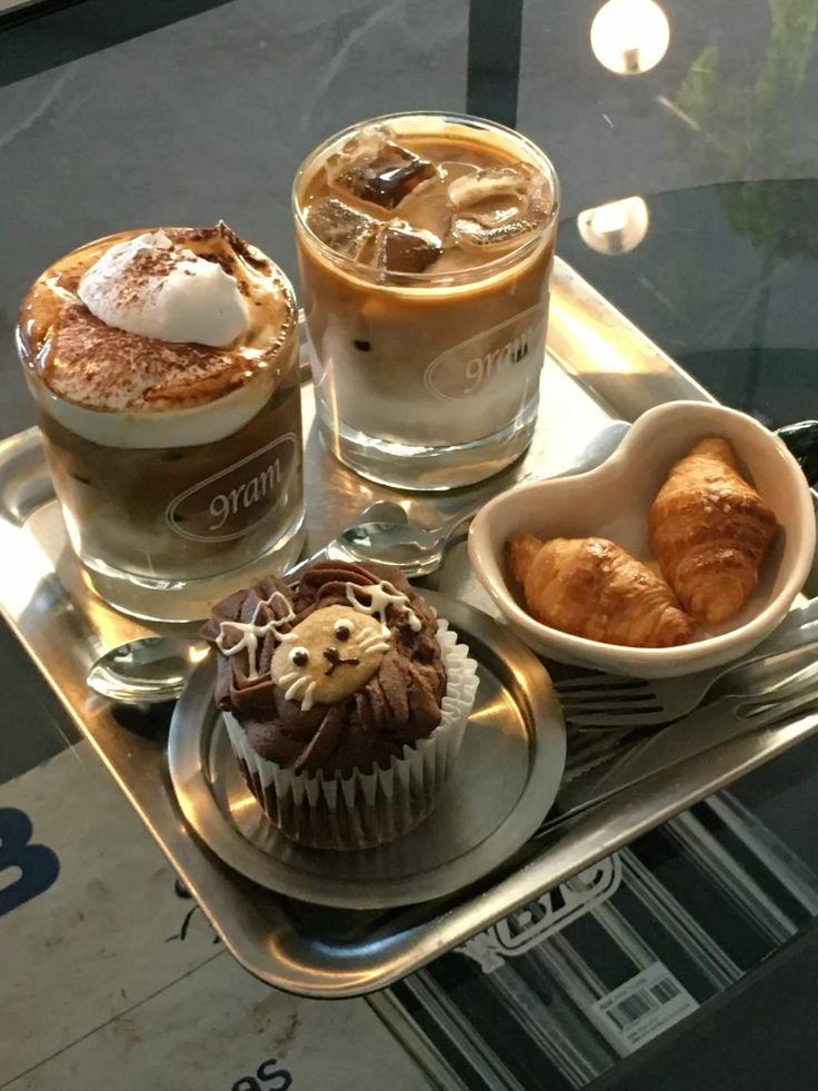 Tray with two coffee drinks, a cat-themed cupcake, and mini croissants, perfect for a delightful coffee break.