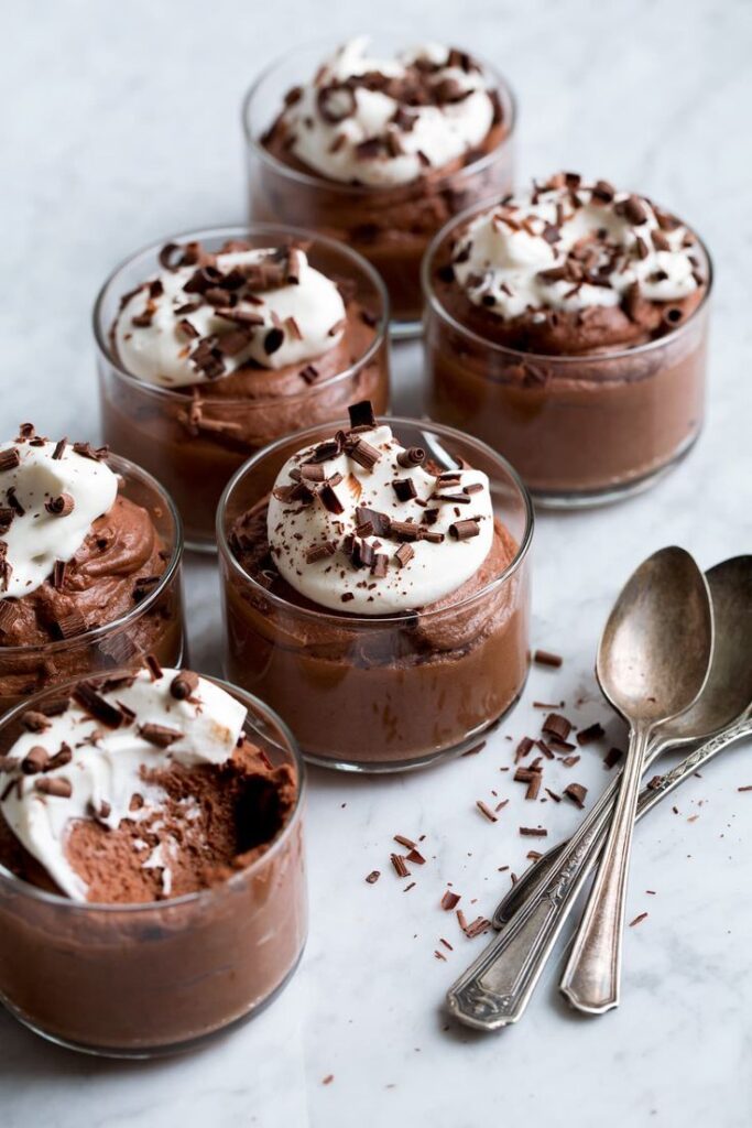 Rich chocolate mousse topped with whipped cream and chocolate shavings in glass cups on a marble surface.