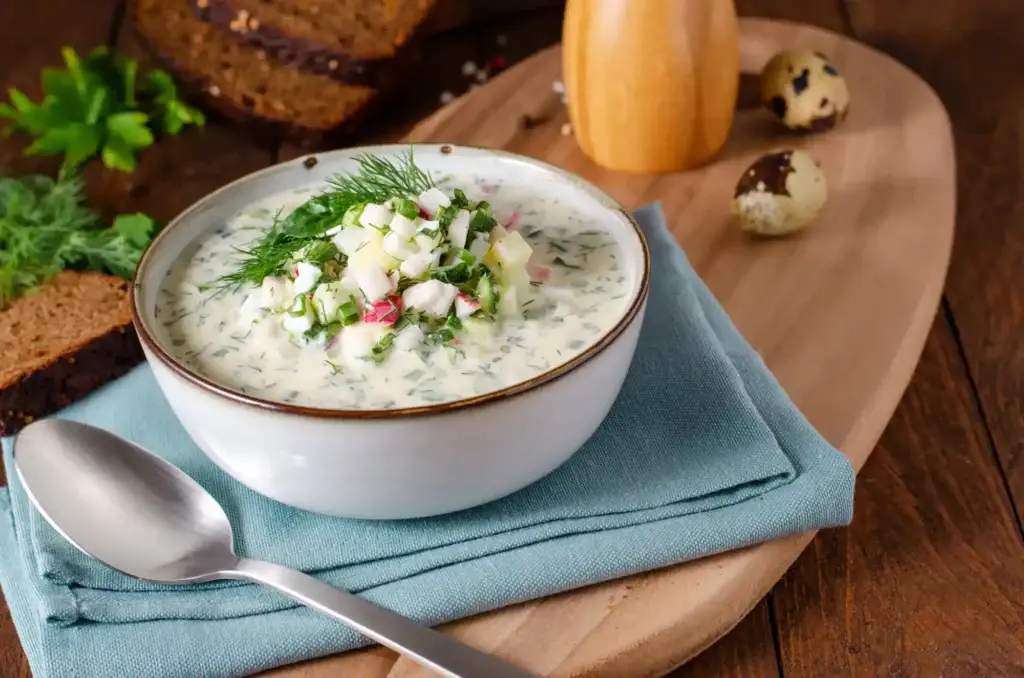 Bowl of creamy cold soup with herbs and diced vegetables on a wooden board, accompanied by bread and quail eggs.