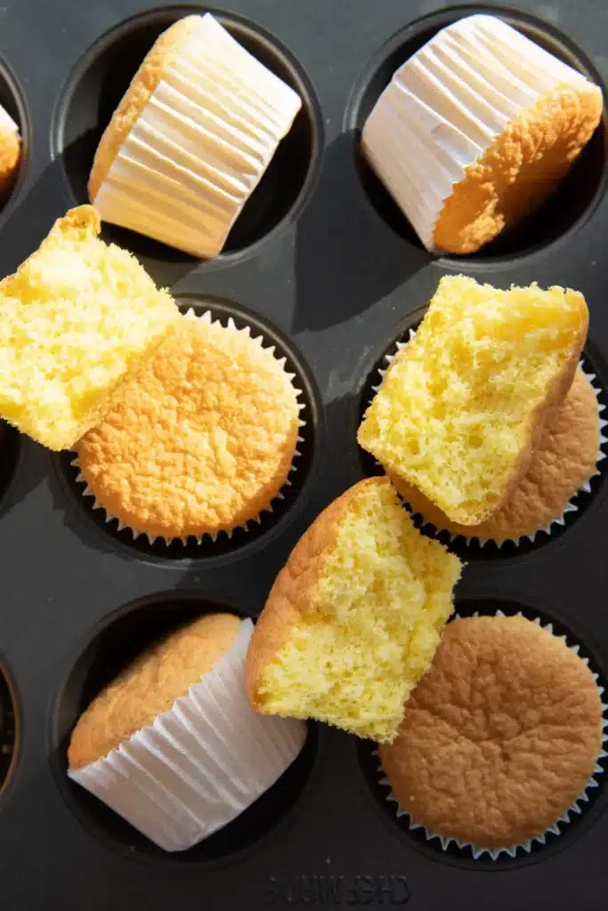 Freshly baked golden cupcakes in a muffin tray, some cut open to show fluffy texture.