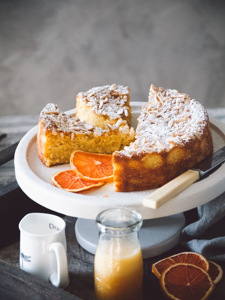 Orange almond cake with coconut flakes on a white stand, garnished with orange slices and juice nearby.