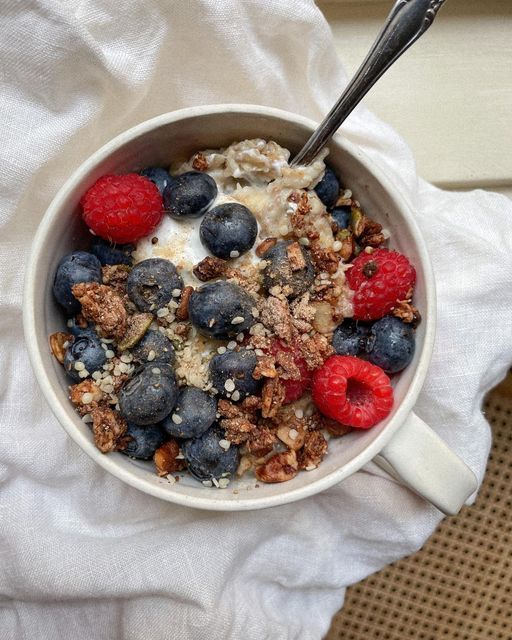 Bowl of oatmeal topped with blueberries, raspberries, granola, and seeds on a white cloth background.