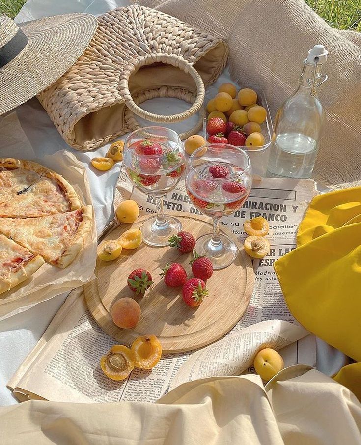 Sunlit picnic setting with fruit, pie, sparkling drinks, and a straw hat on a newspaper-covered blanket.