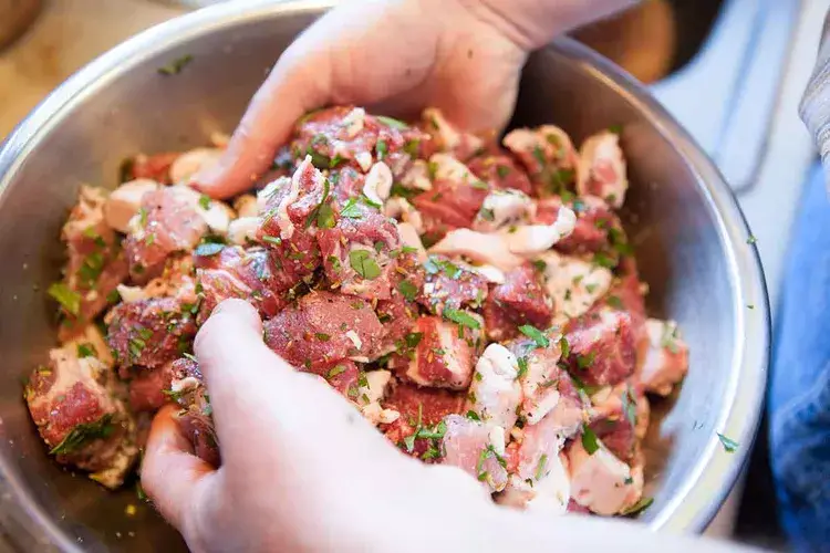 Hands mixing marinated meat cubes with herbs in a bowl, preparing for cooking.