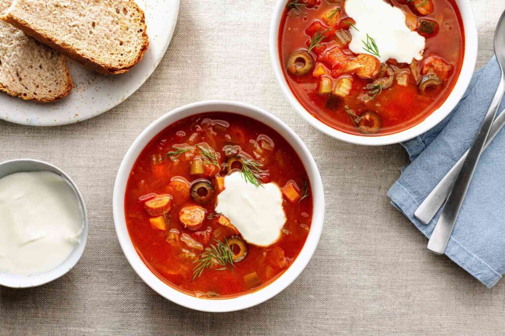 Two bowls of red soup with vegetables and sour cream, served with bread and extra cream on the side.