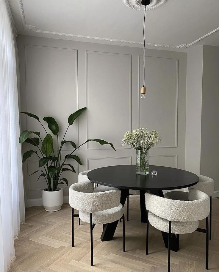 Modern dining room with round black table, white chairs, potted plant, vase of flowers, and minimalist decor.