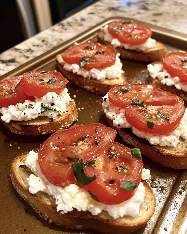 Toasted bruschetta topped with ricotta, sliced tomatoes, fresh herbs, and black pepper on a baking sheet.