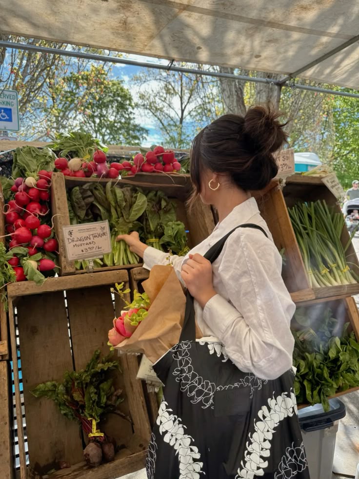 A woman shopping at a farmers market, selecting fresh greens and radishes on a sunny day.