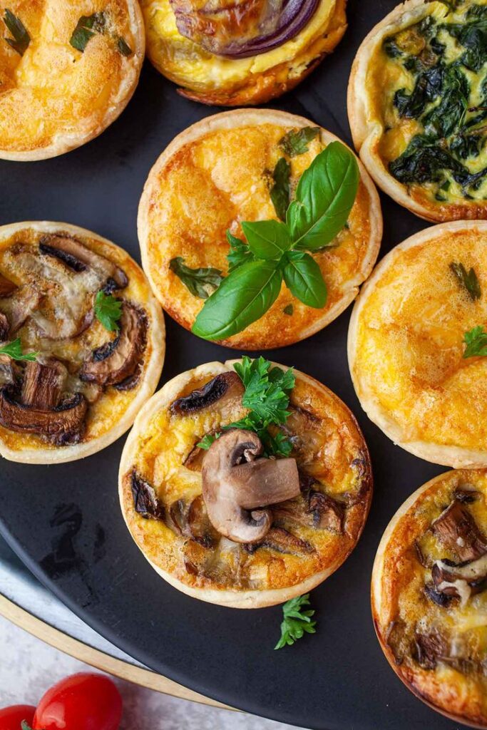 Assorted mini quiches with mushrooms, spinach, and herbs on a black plate, garnished with fresh basil leaves.