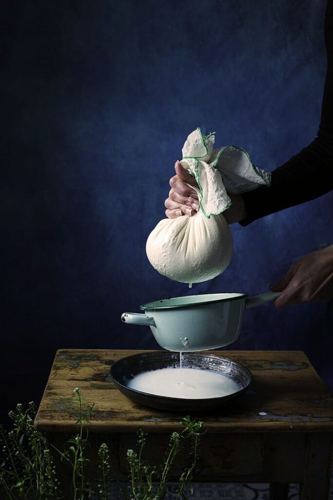 Person squeezing cheesecloth over bowl, making homemade cheese or yogurt, with a dark blue background.