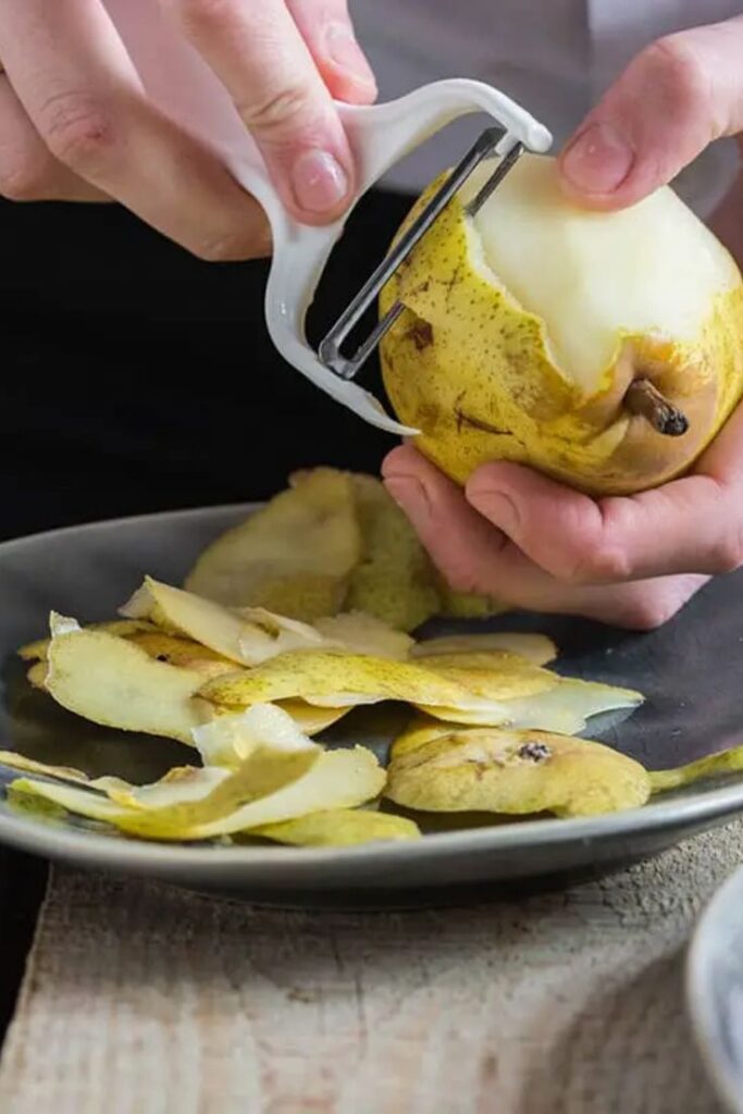 Person peeling a pear with a peeler; fresh pear skin on a plate.