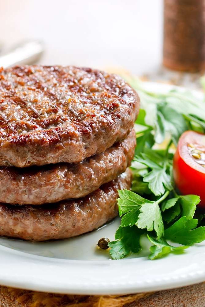 Juicy beef patties stacked on a plate with fresh parsley and a tomato slice. Delicious and savory meal option.