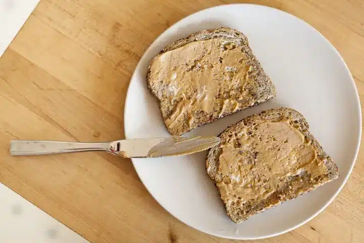 Whole grain bread slices with peanut butter on a white plate, wooden table background, with a butter knife nearby.