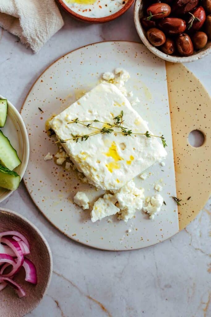 Plate of feta cheese with olive oil and thyme, surrounded by olives, cucumber slices, and red onion on a marble surface.