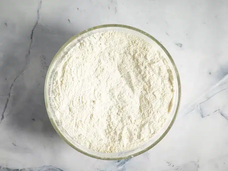 Bowl of white flour on a marble countertop, ideal for baking or cooking recipes, top view close-up.