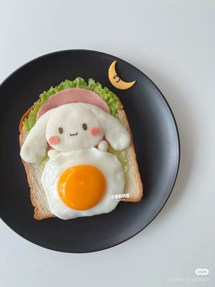 Cute breakfast toast with egg, lettuce, ham, and a smiling face, served on a black plate with a crescent moon garnish.