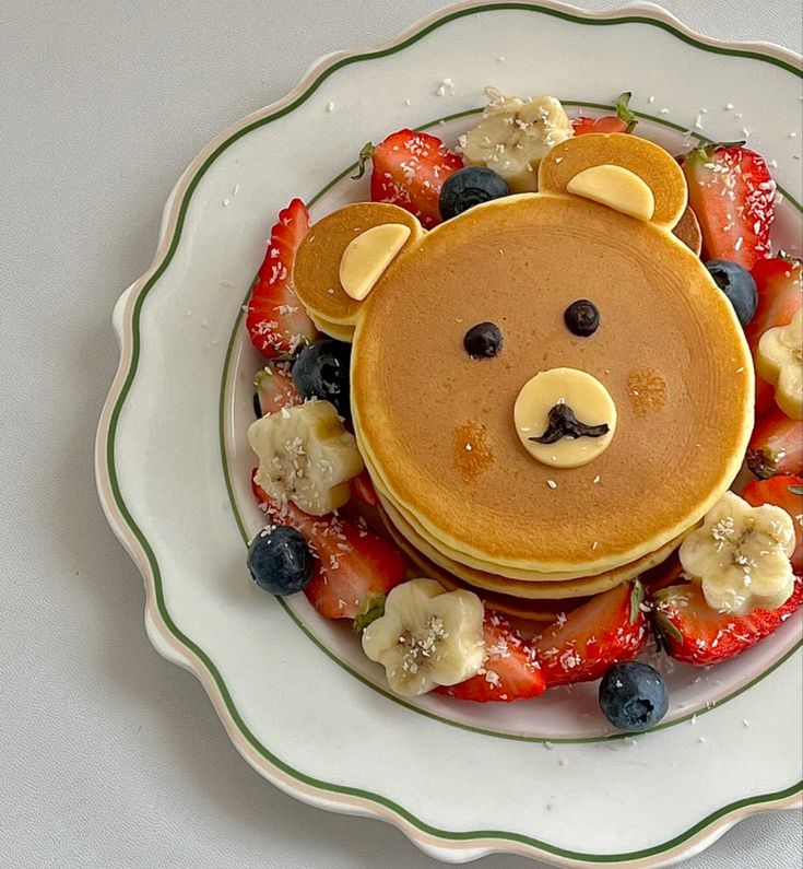 Bear-shaped pancake with fruit garnish, including strawberries, bananas, and blueberries, on a white plate.