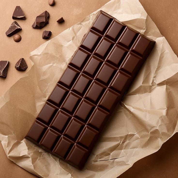 Rich dark chocolate bar on brown paper with chocolate pieces scattered around. Perfect for gourmet dessert lovers.