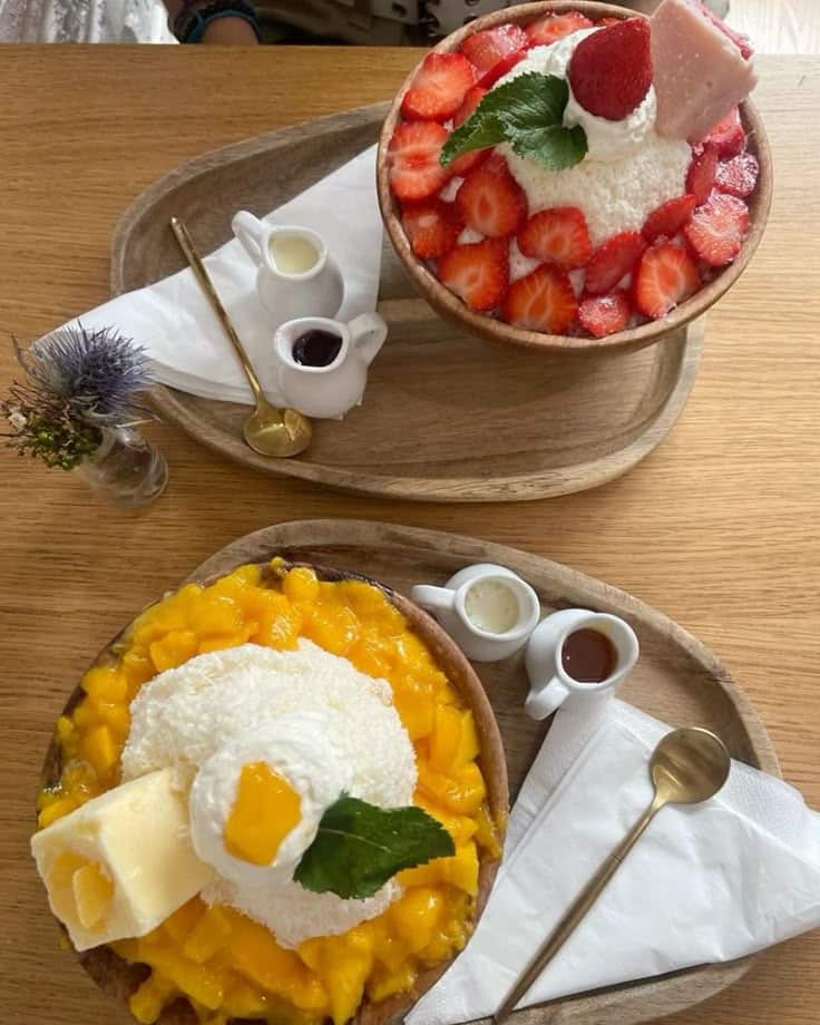 Delicious shaved ice desserts with fresh strawberries and mangoes, topped with cream and syrup on a wooden table.